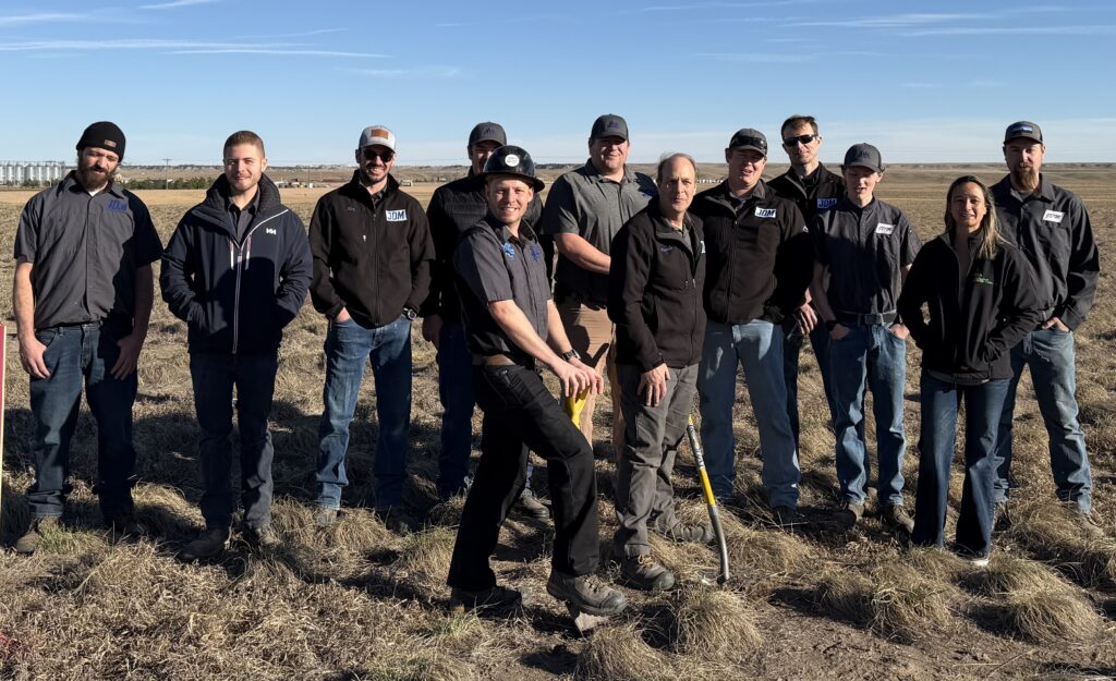 Here is the descriptive text for your groundbreaking photo, designed for SEO and user experience. Alt Text: A group of people, including Rocky Mountain Snacks team members and development partners wearing "JDM" logos, poses for a groundbreaking ceremony. One person in a hard hat holds a shovel, making the first turn of dirt in a vast, grassy field with a distant grain silo.